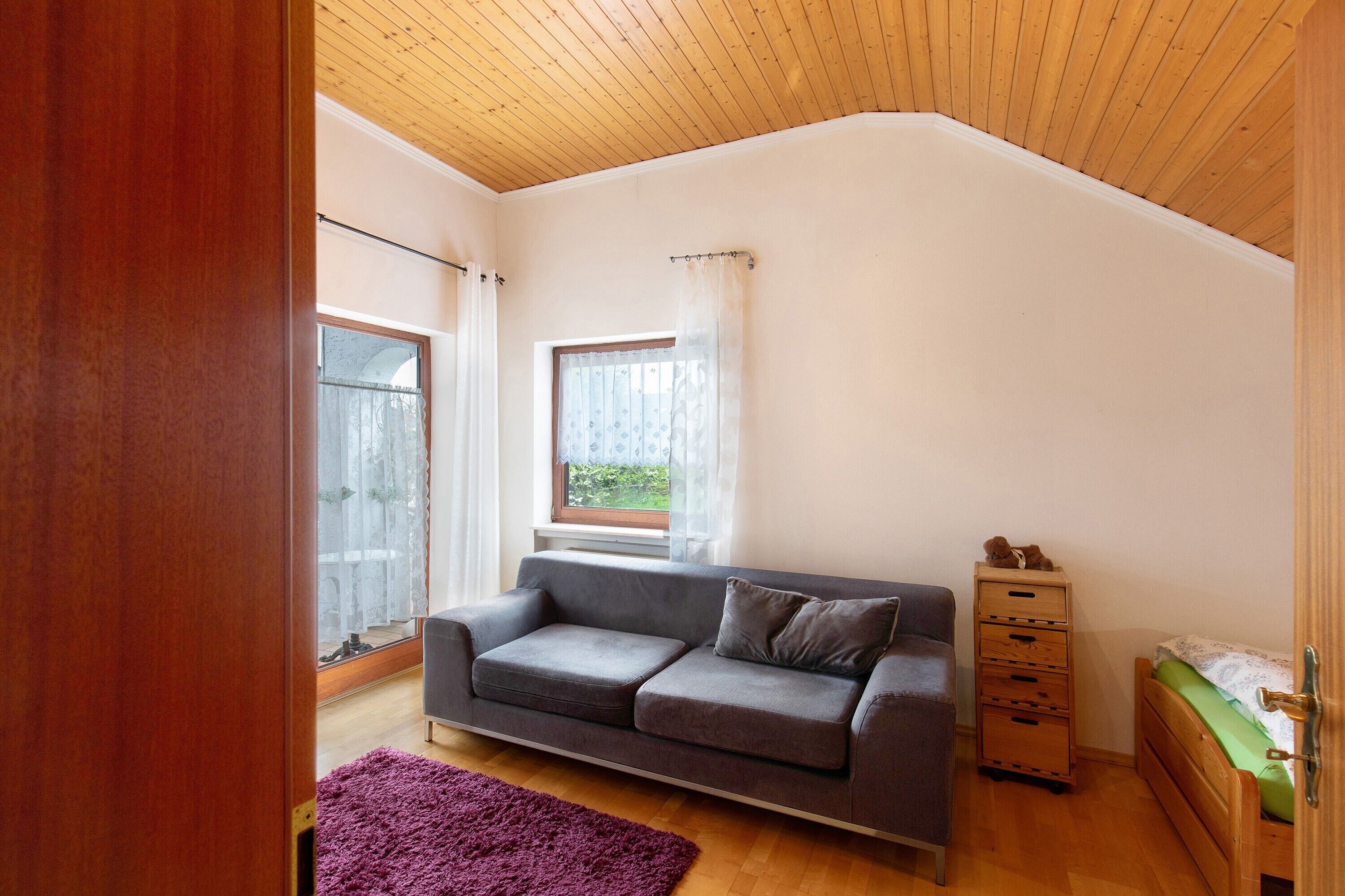 Habitació amb sostre de fusta, un sofà gris, una petita cuixa de fusta, un llit amb matalàs verd i una catifa morada.
