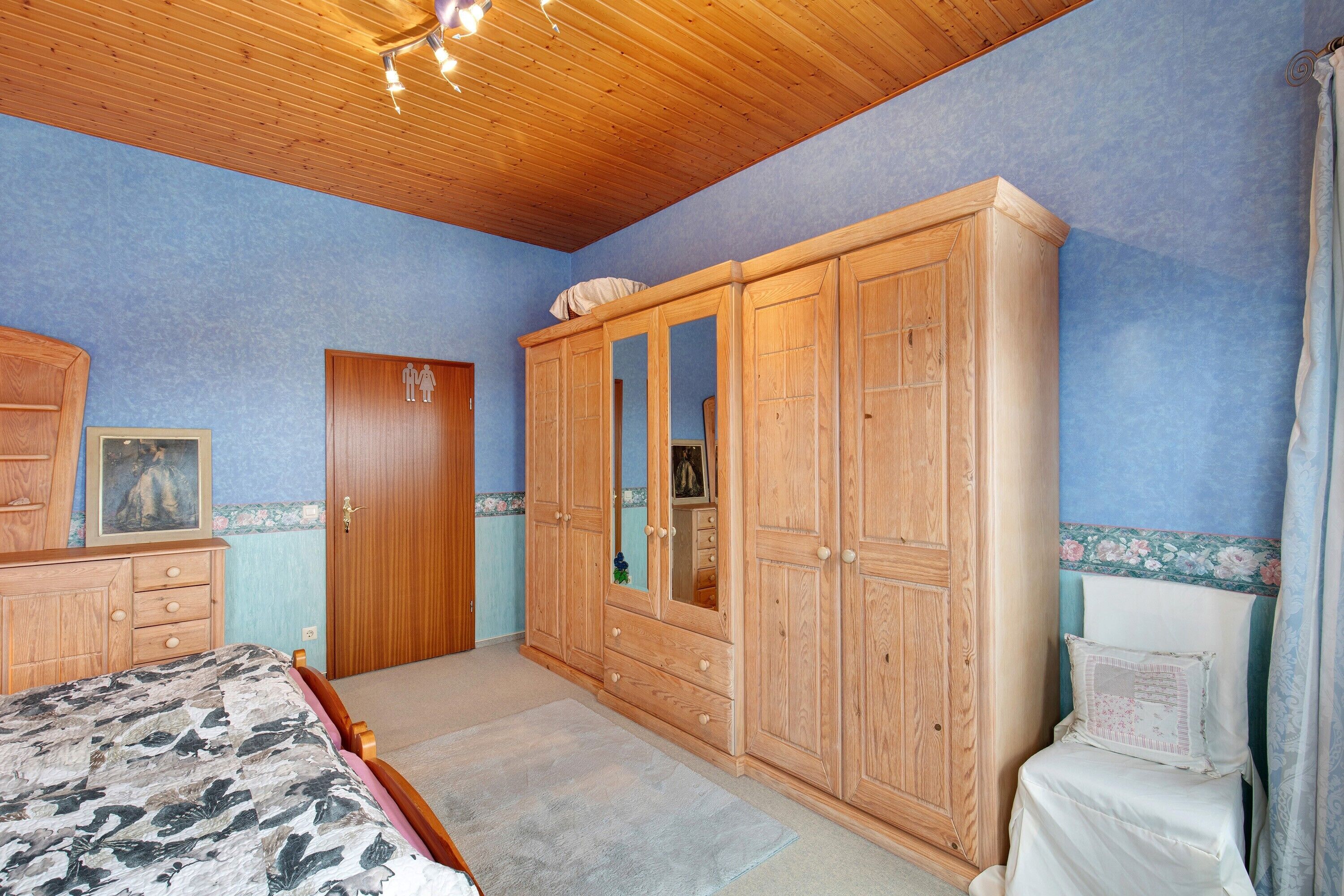 A bedroom with a wooden wardrobe, a bed with a patterned bedspread, and a small bedside table.