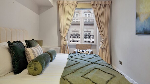 Une chambre à coucher avec un lit, des oreillers, une fenêtre avec des rideaux, une chaise et une œuvre d’art accrochée au mur.