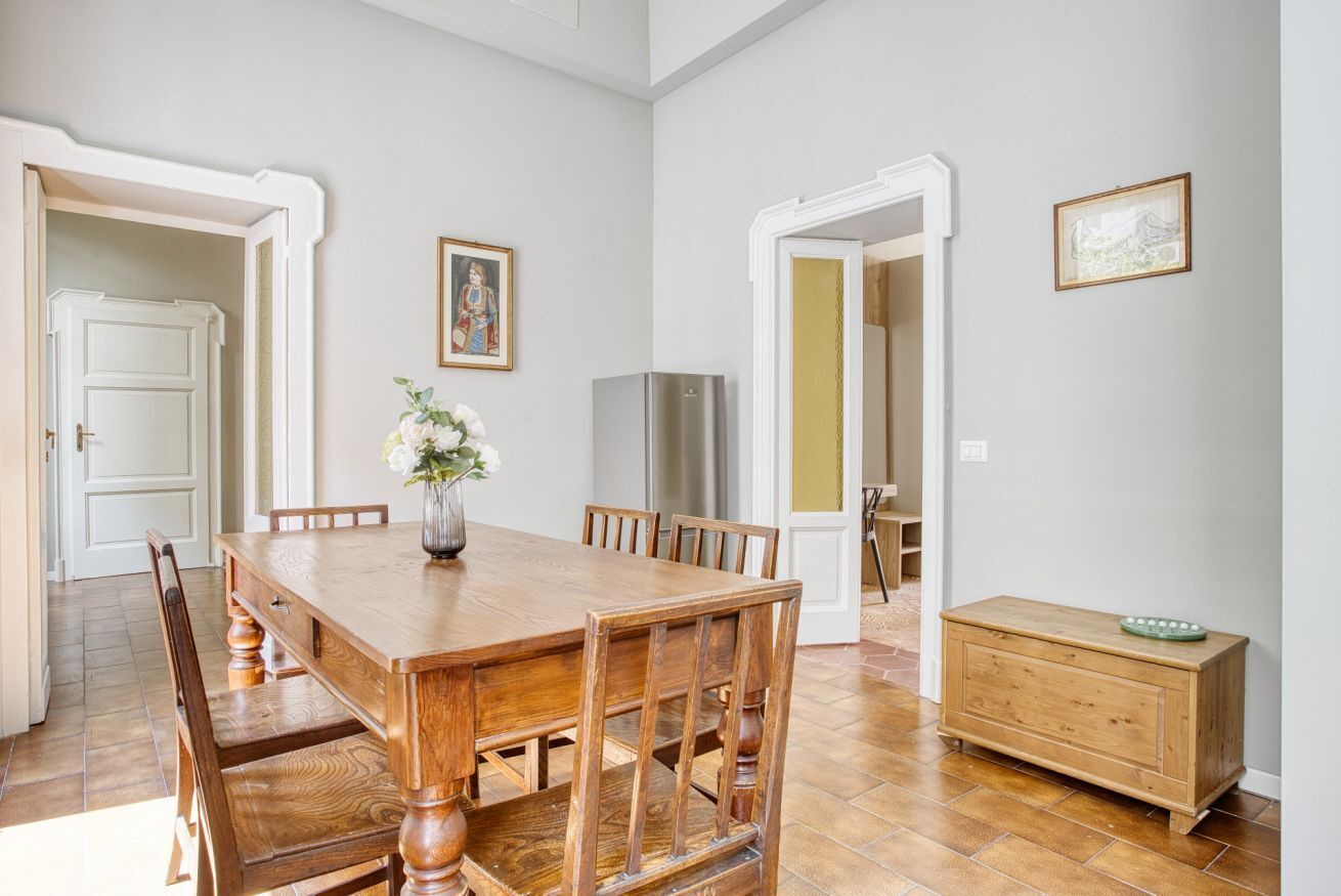Un comedor con una mesa y sillas de madera, un jarrón con flores, un refrigerador y un armario de madera.
