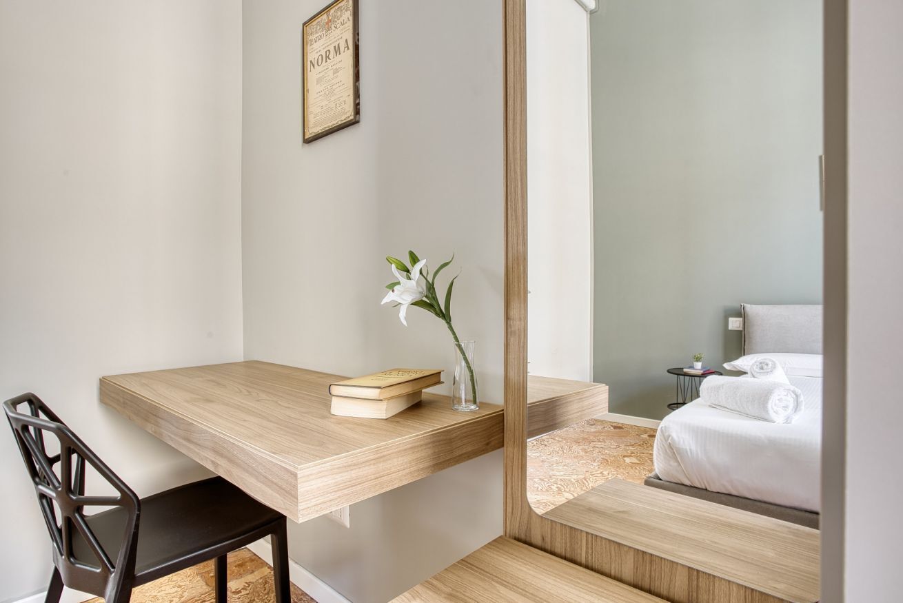 A modern room with a wooden desk, a chair, a bedside table with a book and a vase of flowers, and a bed with neatly folded towels.