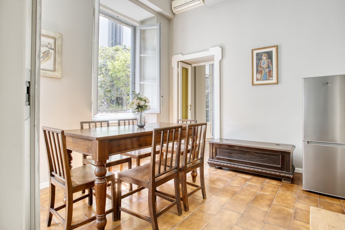 A dining area with a wooden table and chairs, a window with a view of greenery, a refrigerator, and a cabinet.