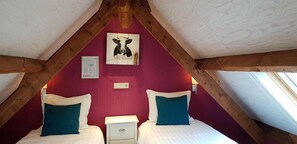 Ein raumförmiger Raum mit Holzbalkendecke, zwei Betten mit weißer und türkisfarbener Bettwäsche, einem an der Wand montierten Lichtschalter und einem gerahmten Bild von einer Kuh.
