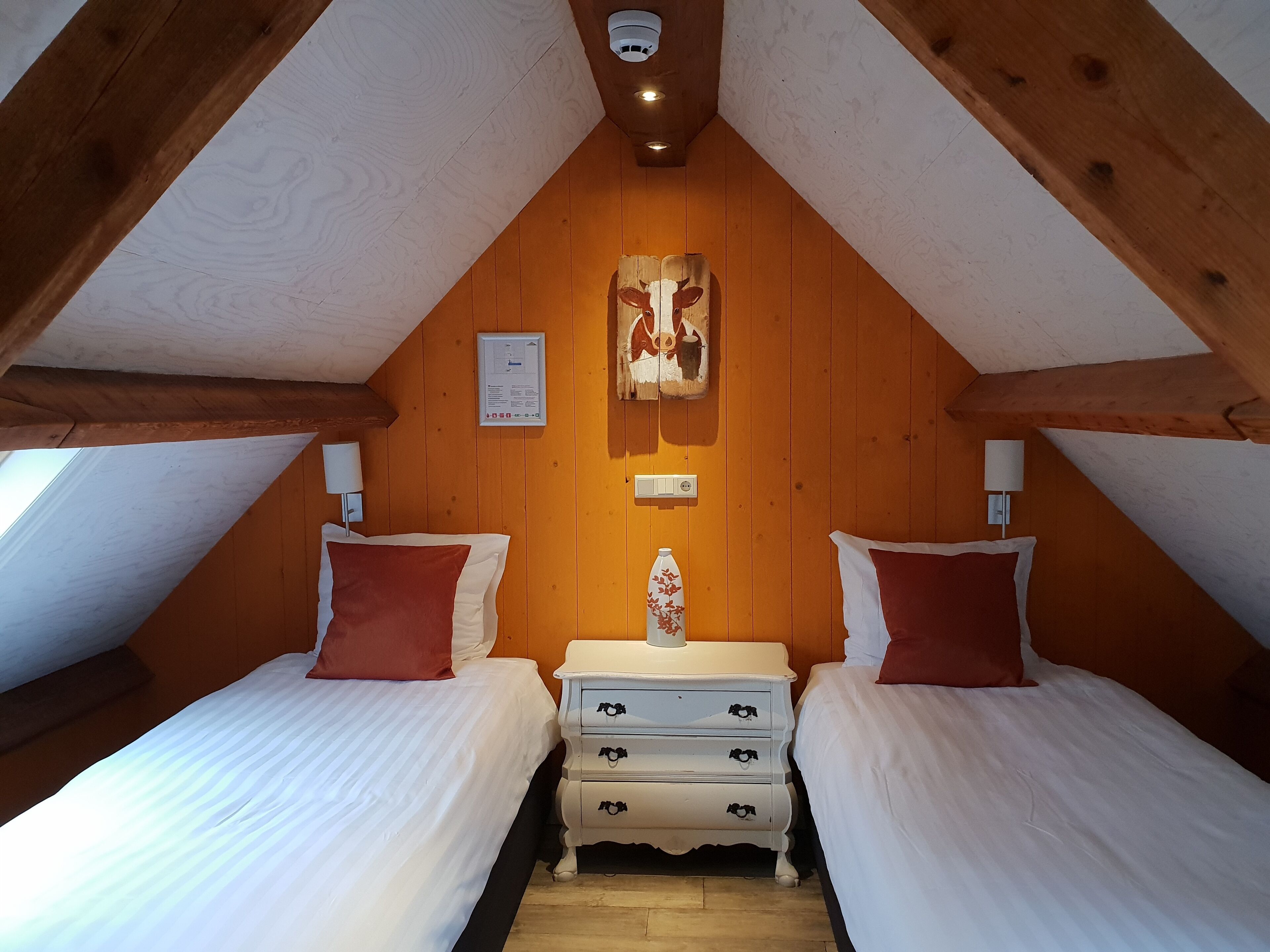 Een knus zolderkamer met twee bedden, houten balken, een plafondlamp en een wandlamp.