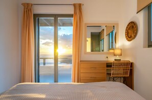 A bedroom with a bed, a desk, a chair, a mirror, and a view of the sunset through a sliding door. - Phos Villas Tinos (Tinos)