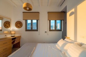 A bedroom with a bed, a desk, two windows with blinds, a wall-mounted lamp, and a ceiling light fixture.