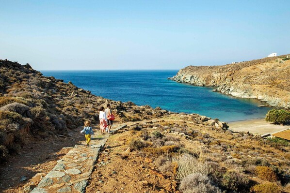 Camino costero rocoso que conduce a una playa con mar azul cristalino y algunos edificios al fondo.