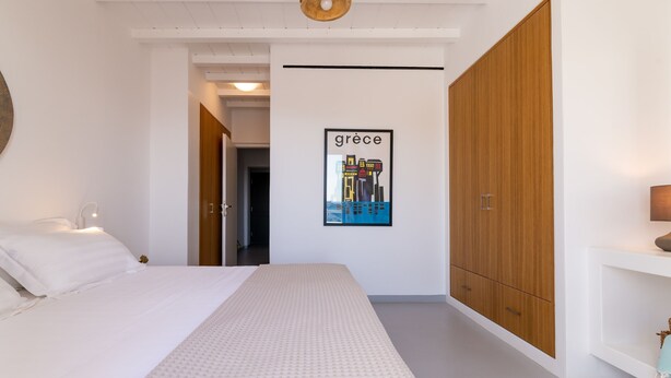 A bedroom with a bed, a wooden wardrobe, a white wall with a framed picture, and a door leading to another room.