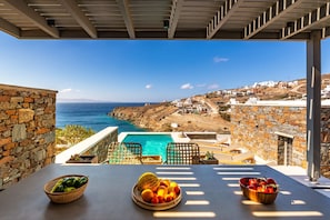 A terrace with a pool, stone walls, and a view of the sea and hillside buildings. - Phos Villas Tinos (Tinos)