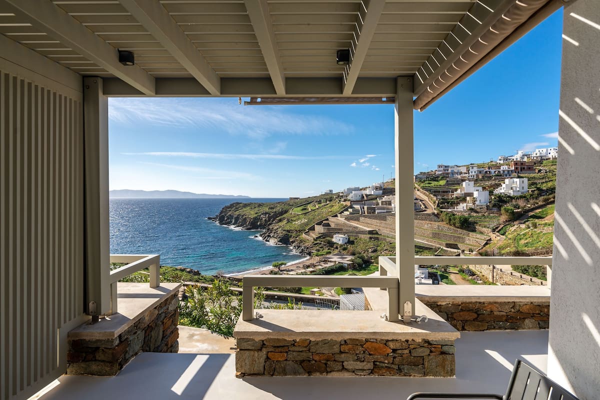 A balcony with a stone ledge, a view of the ocean, and buildings on a hillside.