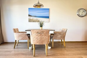 Een eethoek met een tafel en stoelen, een klok aan de muur en een schilderij van een zandstrand boven de tafel.