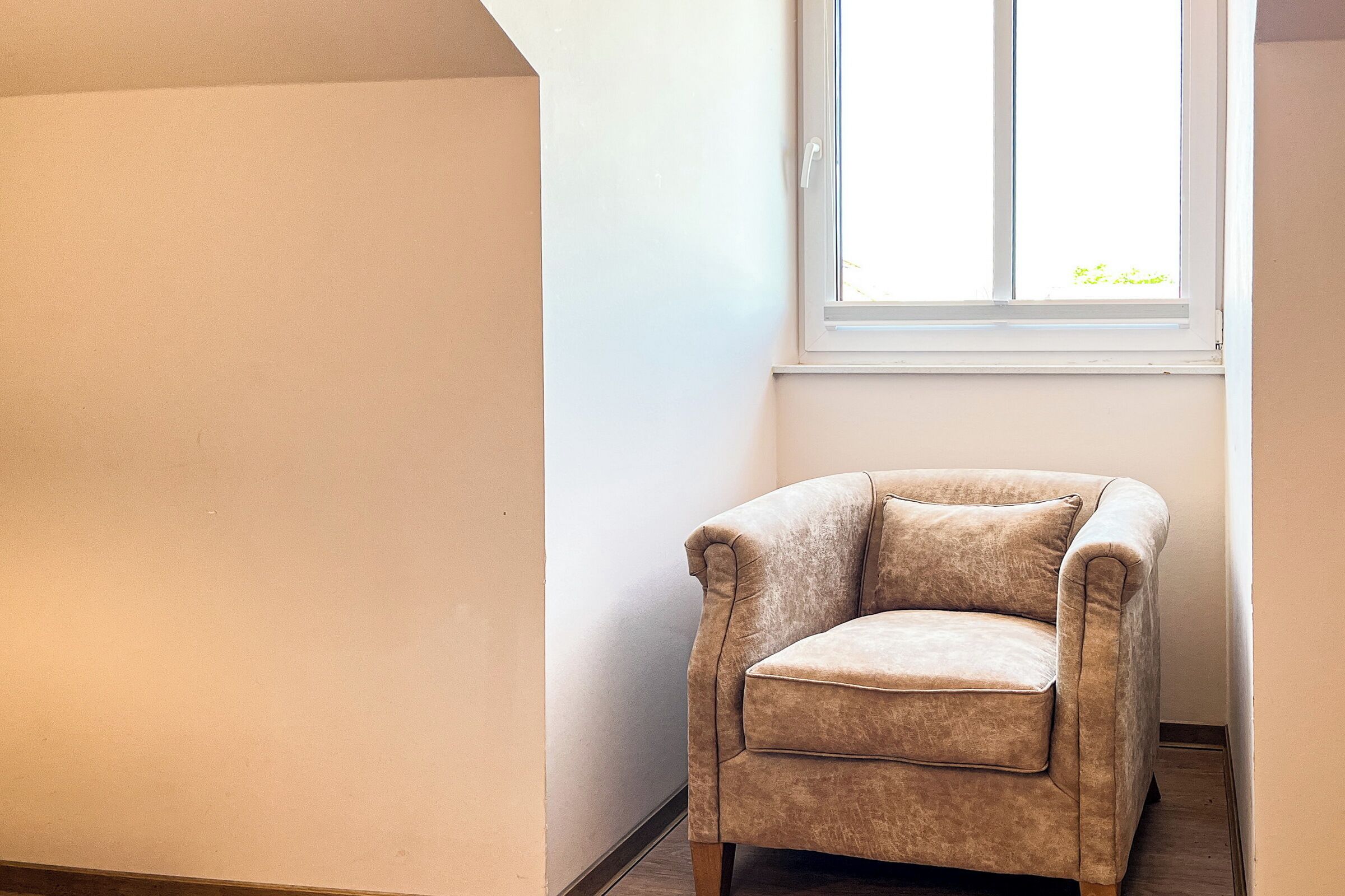 A corner room with a beige armchair, a window, and a plain wall.