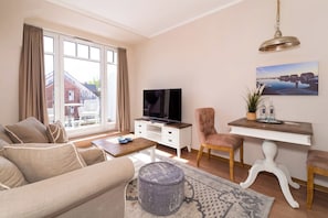 Een woonkamer met een bank, voetenbankje, salontafel, televisie, eettafel, stoel en een schilderij aan de muur.