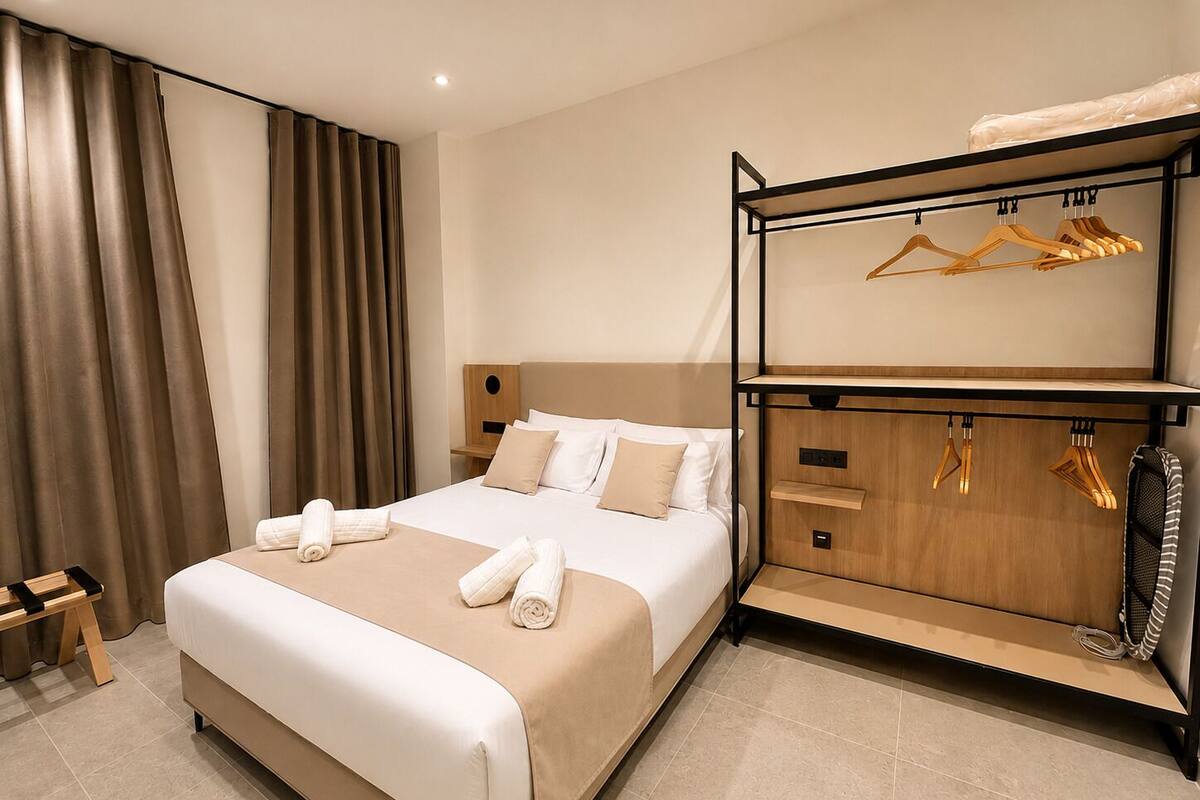 A hotel room with a bed, a wooden bench, a coat rack with hangers, and a built-in wardrobe.