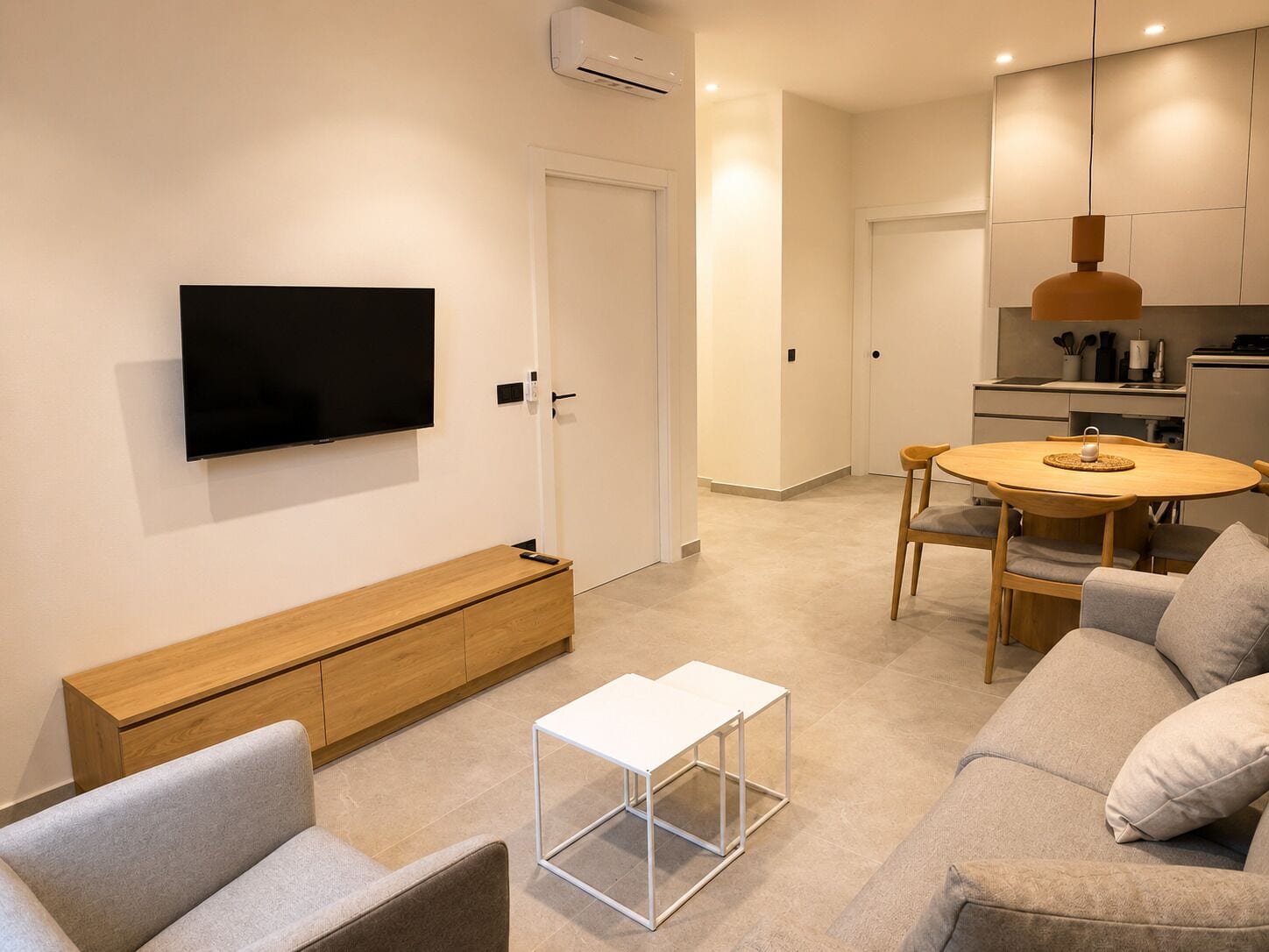 A modern living room with a flat-screen TV, a wooden bench, a white coffee table, a grey sofa, and a dining area with a round table and chairs.