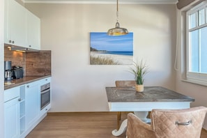 Eine kompakte Küche mit weißen Schränken, einem eingebauten Backofen und einer Kaffeemaschine. Ein Esstisch mit einer Zimmerpflanze und einem Strandbild darüber.