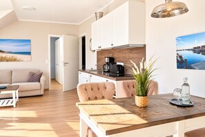 Una cocina moderna con armarios blancos, encimera de madera y una cafetera. Hay una mesa de comedor con sillas y una planta en maceta.