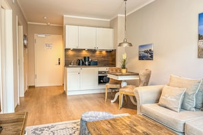 Eine moderne Küche mit weißen Schränken, ein Essbereich mit Holztisch und -stühlen sowie ein Wohnzimmer mit beiger Couch und Couchtisch.