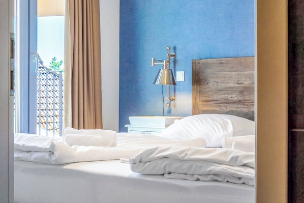 Ein Hotelzimmer mit Bett, Nachttisch, Lampe und Blick auf einen Balkon mit Vorhängen.
