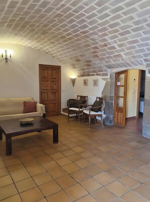 Ein Wohnzimmer mit einem weißen Sofa, einem Holztisch, zwei Korbstühlen, einem gefliesten Boden, einer Ziegeldecke und einer Holztür.
