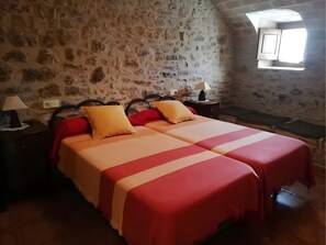 Ein Zimmer mit Steinwand, einem Bett mit rot-weißer Bettwäsche, einem Nachttisch mit Lampe und einem Fenster mit Tageslicht.