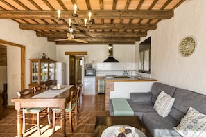 Ein Essbereich mit Holztisch und -stühlen, eine Küche mit Herd und Backofen sowie ein graues Sofa mit gemusterten Kissen.