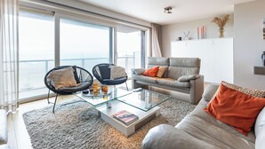 Una sala de estar moderna con una mesa de centro de vidrio, dos sillones, un sofá y vistas al mar a través de amplios ventanales.