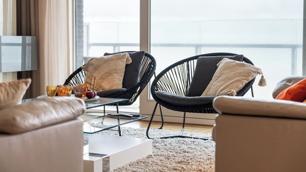 Un salón moderno con dos sillones de mimbre negro, una mesa de centro de vidrio y un sofá beige.