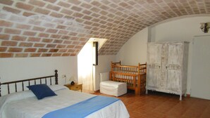 Un dormitori amb sostre de maó, un llit amb almofada blava, una bressol de fusta i un armari blanc.