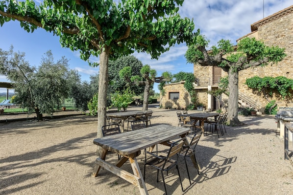 An outdoor patio with wooden tables and chairs, surrounded by trees and a stone building.