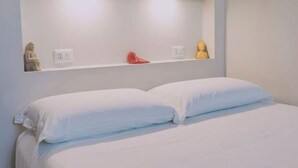 A bedroom with a bed, white pillows, a shelf with decorative items, and a wall socket. - Apartment with panoramic terrace in historic center (Napoli)