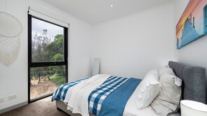 A bedroom with a large window, a bed with a checkered blanket, a grey headboard, and a painting on the wall. - Beachfront Gem in Capel Sound (Capel Sound)