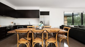 A modern kitchen with dark cabinetry, a dining table with wooden chairs, and a black sofa in a bright room with large windows. - Beachfront Gem in Capel Sound (Capel Sound)