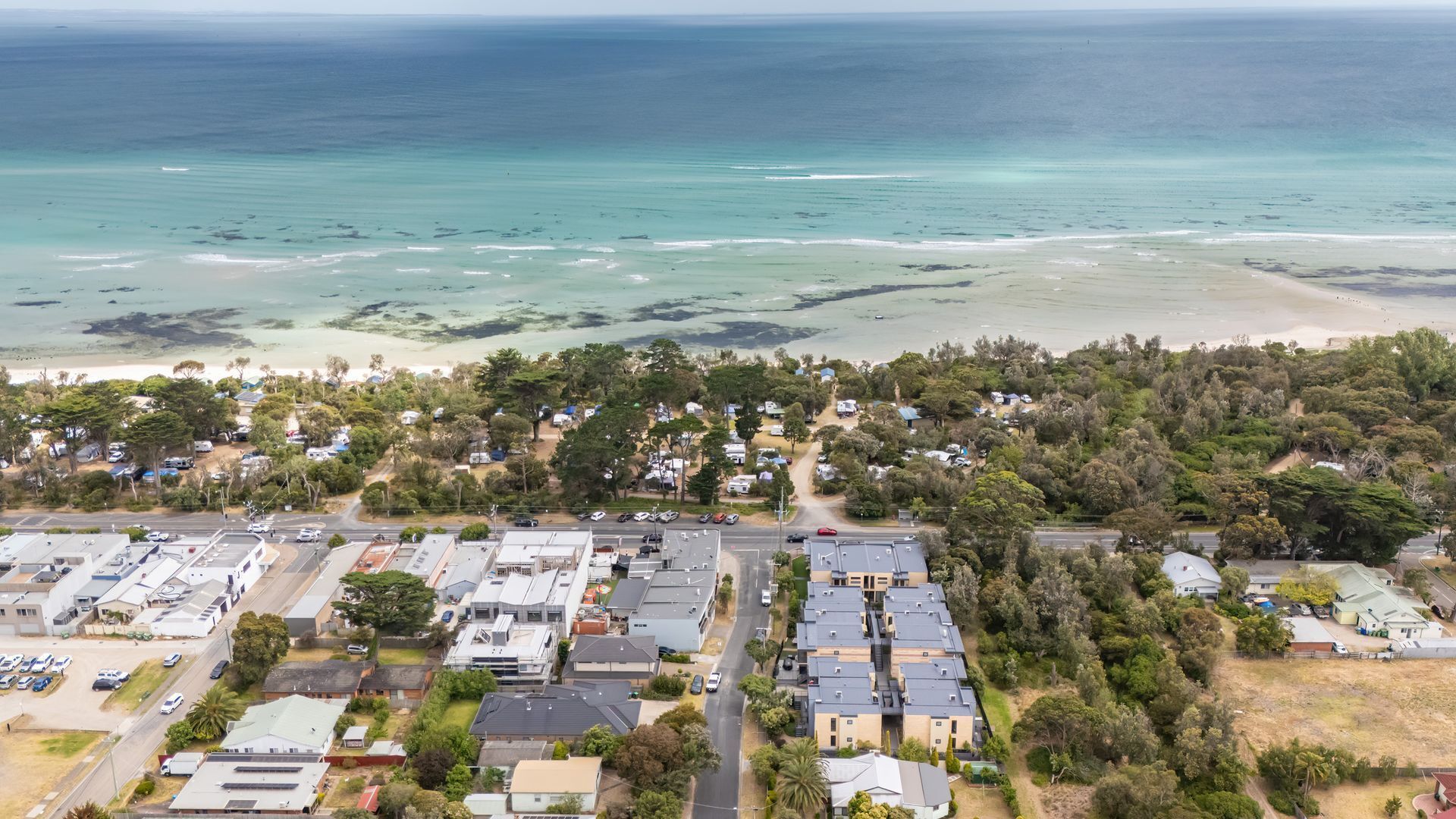 An aerial view of a coastal residential area with houses, a beach, and a road.