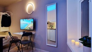 A modern room with a flat-screen TV mounted on the wall, a small desk with a chair, a large mirror, and a window with candles. - Cozy, Central Studio by the Bus Station (Bergen)