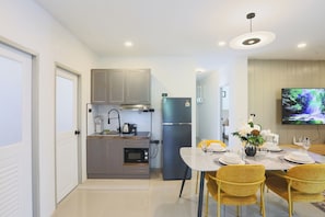 Eine moderne Küche mit grauen Schränken, einem eingebauten Backofen und einem schwarzen Kühlschrank. Es gibt einen Essbereich mit einem weißen Tisch, gelben Stühlen und einem an der Wand befestigten Fernseher.