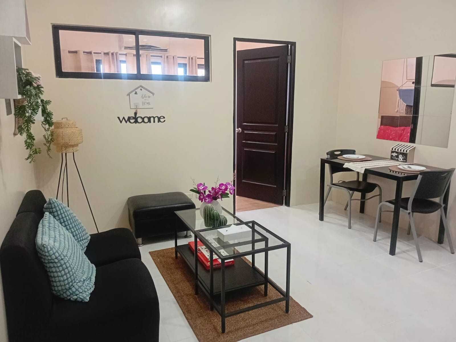 A living room with a black sofa, a glass coffee table, a small side table, and a dining area with a table and chairs. A 'Welcome' sign is on the wall, and a door is visible.