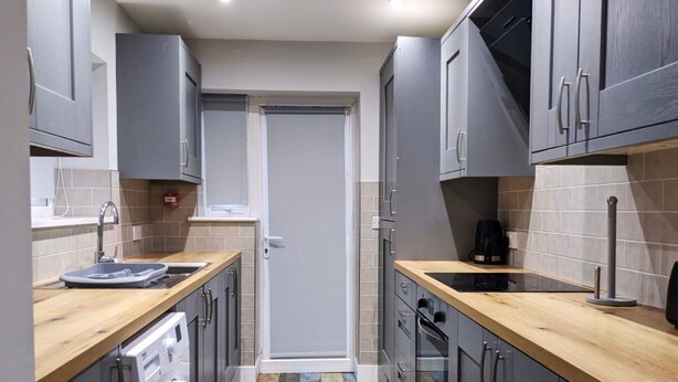 A compact kitchen with grey cabinets, a wooden countertop, a washing machine, and a coffee maker. - The Red Lounge House - sleeps 14 (Liverpool)