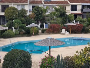 Une piscine dotée de parasols, entourée de verdure et bordée d’une rangée de bâtiments aux toits couverts de tuiles.