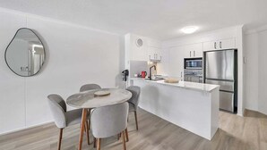 A modern kitchen with white cabinetry, a stainless steel refrigerator, and a round dining table with grey chairs. - Alex Beach Stay | Pool, Gym, Beachfront walks (Sunshine Coast)