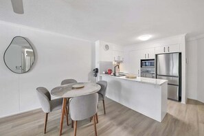 A modern kitchen with white cabinetry, a stainless steel refrigerator, and a round dining table with grey chairs. - Alex Beach Stay | Pool, Gym, Beachfront walks (Sunshine Coast)