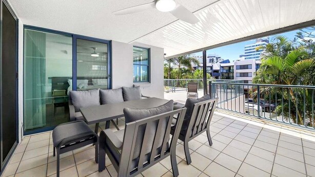 A balcony with a dining table and chairs, a ceiling fan, and a view of palm trees and buildings. - Alex Beach Stay | Pool, Gym, Beachfront walks (Sunshine Coast)