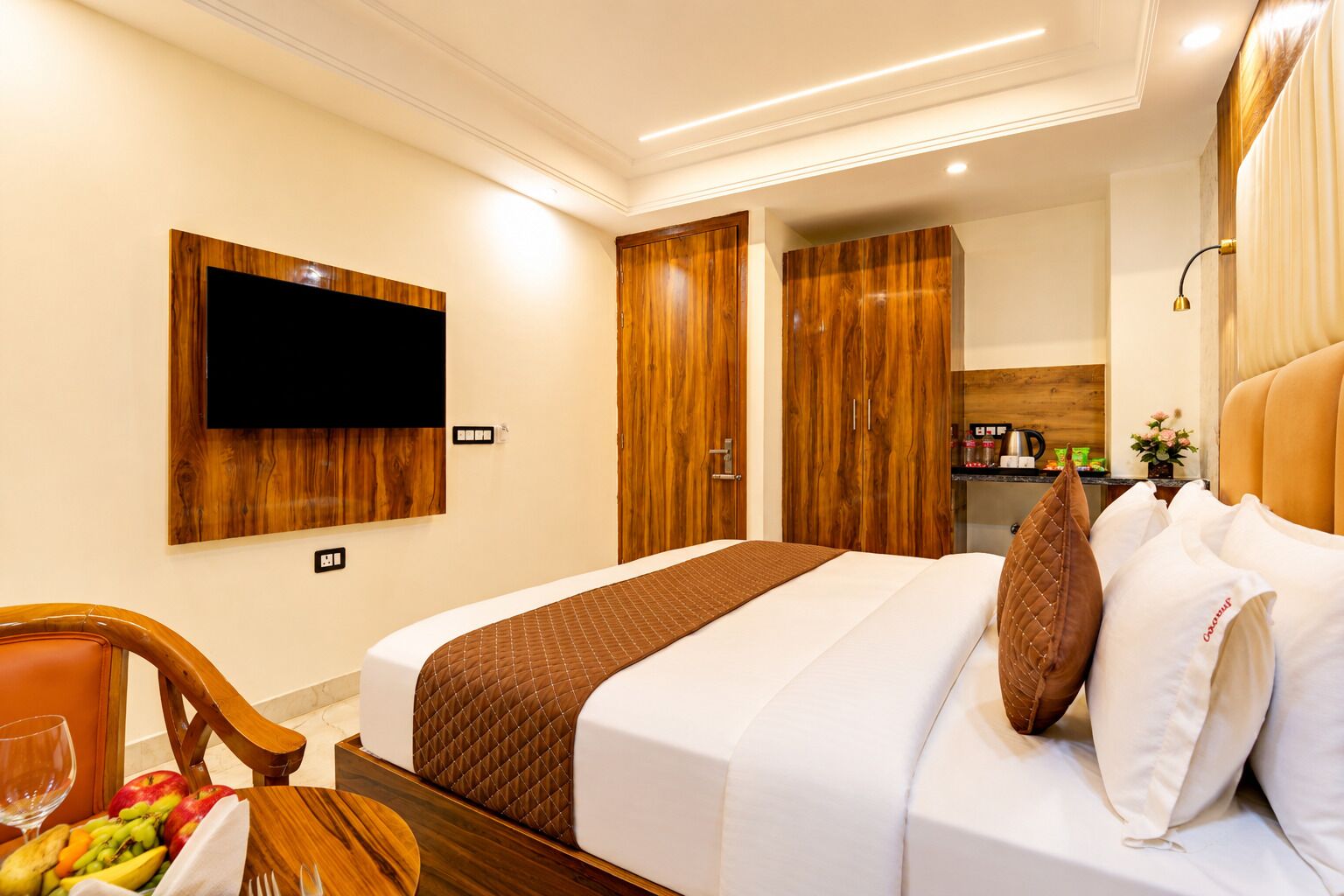 A hotel room with a bed, a TV mounted on the wall, wooden wardrobes, a small kitchenette, and a dining area with a table and fruit.