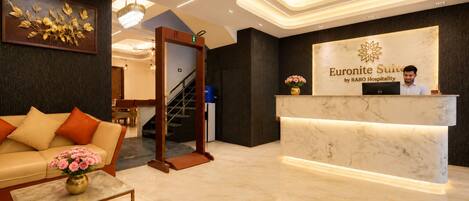 A hotel reception area with a marble reception desk, a sofa with cushions, a coffee table with a vase, and a staircase leading to another floor.