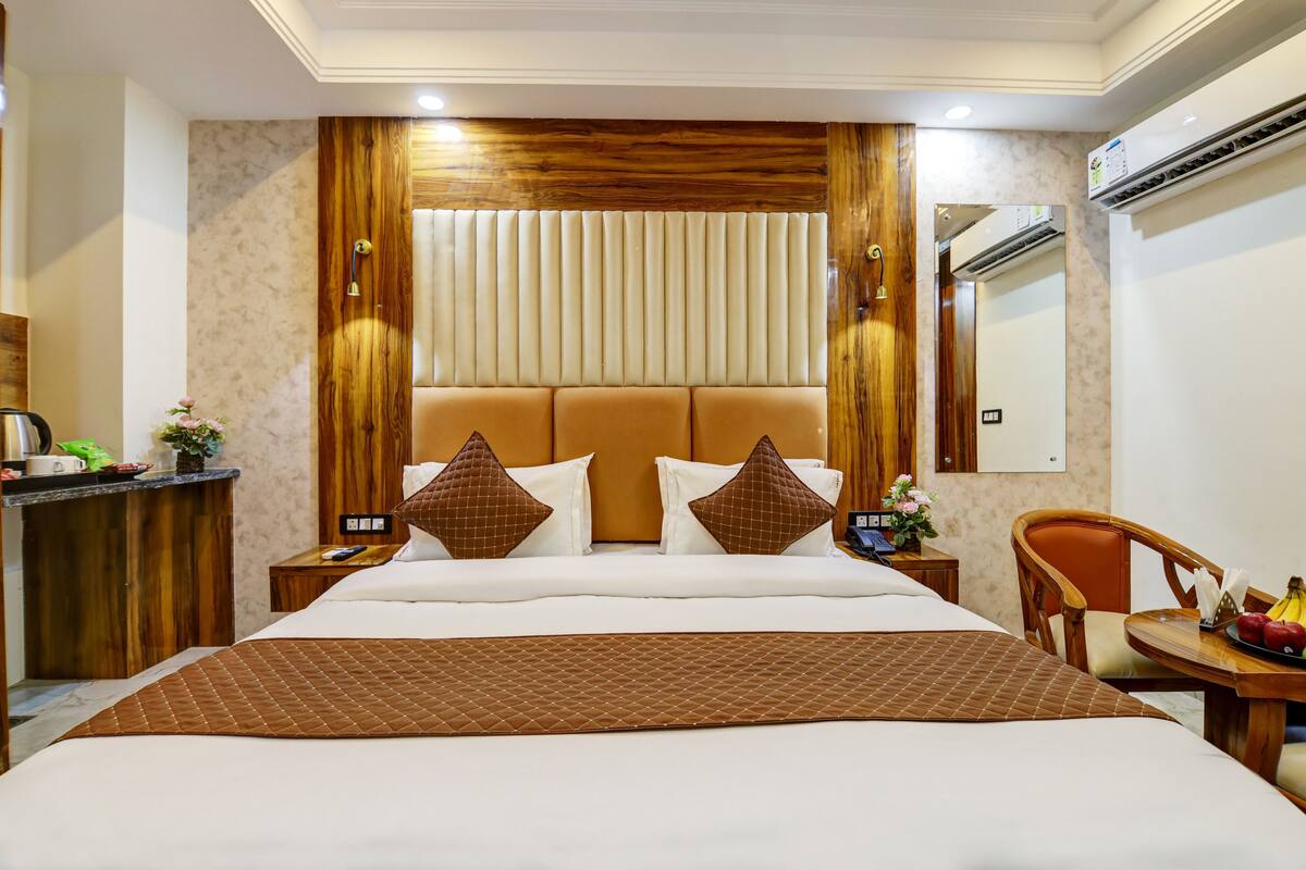 Habitación de hotel con una cama grande, cabecera de madera, dos mesitas de noche, una pequeña mesa con fruta y un equipo de aire acondicionado.