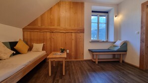 Et hyggeligt rum med træbjælker i skråvæggen, en indbygget bænk med hynder, et lille bord med en plante og et vindue med udsigt til træer.