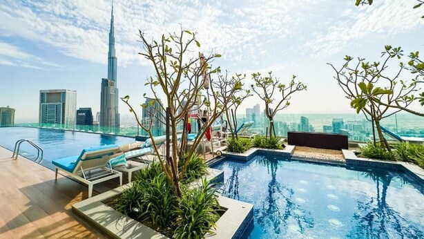 A rooftop infinity pool with lounge chairs, surrounded by greenery, overlooking a city skyline with a prominent skyscraper. - Lucky Getaways (Dubai)