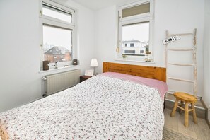 A bedroom with a floral bedspread, two windows, a wooden ladder, a small stool, and a bedside table with a lamp. - Vacation apartment Seebrise am Ostdeich (Büsum)