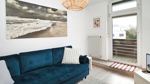 Een moderne woonkamer met een blauwe bank, een grote foto van een strand en een rieten plafondlamp, met een schuifdeur van glas naar een buitenruimte.