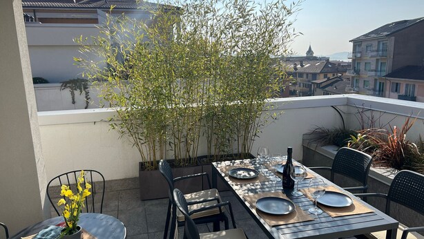 Una terraza en la azotea con una mesa de comedor preparada para dos personas, rodeada de sillas, una planta en maceta y vistas de edificios al fondo.